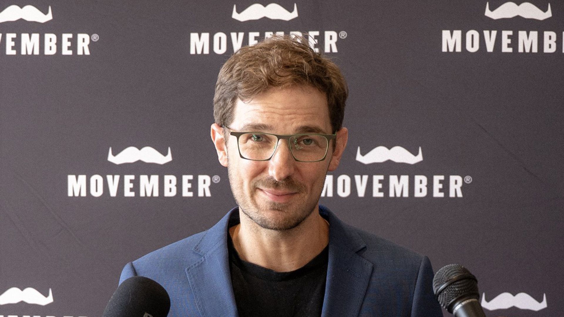 Photo showing Movember staff member in front of a board bearing the Movember moustache logo.