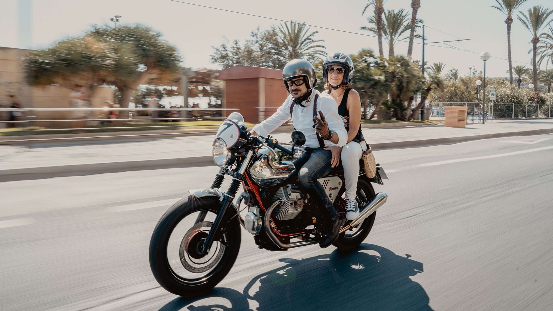 Dapper man riding a classic motorcycle down palm-lined road.